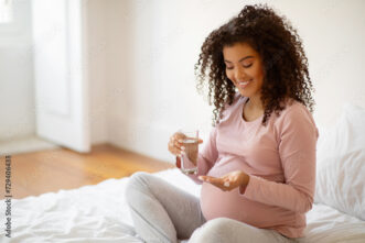 Pregnant person sitting on a bed holding a glass of water and a prenatal supplement, illustrating the importance of folic acid during pregnancy.