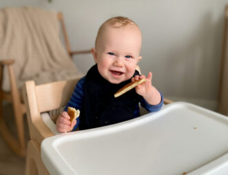 A baby sits in a high chair, smiling while holding pieces of food in both hands.
