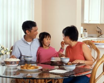 An Asian family, an adult male and female are seated around a table eating a meal with a young female standing in between the adults. National Cancer Institute on Unsplash https://unsplash.com/photos/BQPi8F_UON0