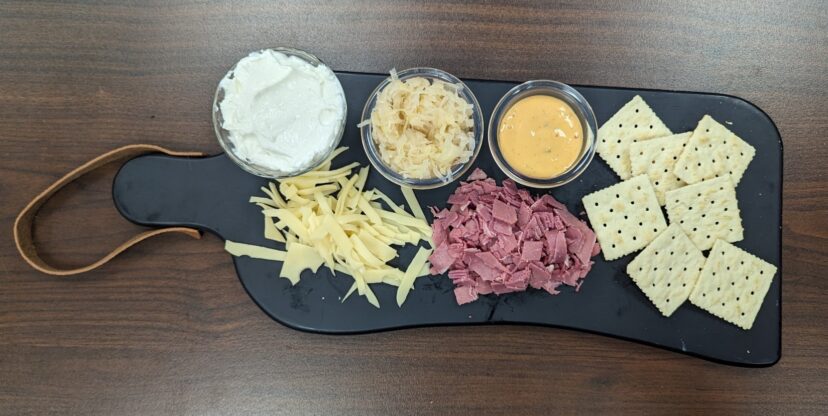 Black rectangle cutting board with small clear cups of ingredients along the top: Sour cream, sour kraut, 1000 island dressing, portions of shredded cheese, and corned beef in front of the cups and saltine crackers displayed next to the ingredients.