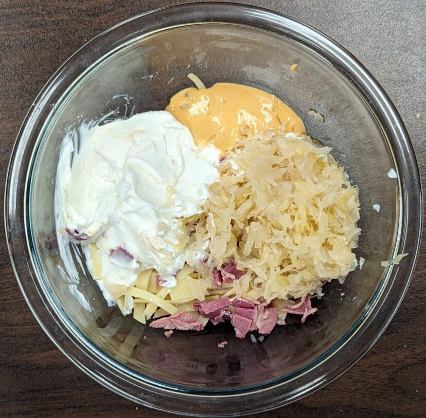 Clear bowl with portions of sour cream, 1000 island dressing, sour kraut, sliced corned beef, and sliced cheese prior to mixing and baking.