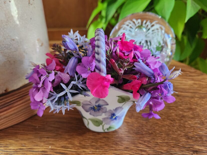 A small china basket arrangement contains salvia, amsonia, Spanish bluebell, and dollar plant flowers.