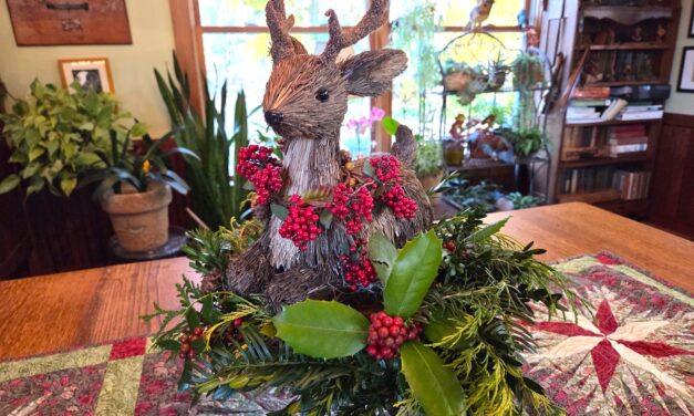 “Twig and straw reindeer figure placed beside a pedestal greenery arrangement.”