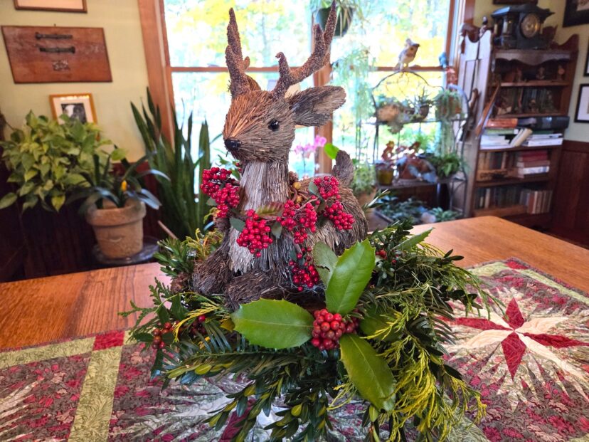 “Twig and straw reindeer figure placed beside a pedestal greenery arrangement.”