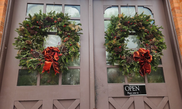“Two large evergreen holiday wreaths decorated with pinecones, berries, and red velvet bows hanging on double glass doors with an ‘Open’ sign.”