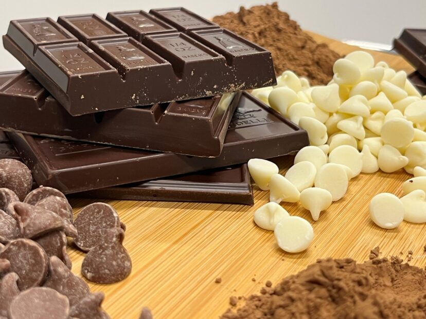 Assorted chocolate types including stacked dark chocolate bars, milk chocolate chips, white chocolate chips, and cocoa powder on a wooden cutting board.