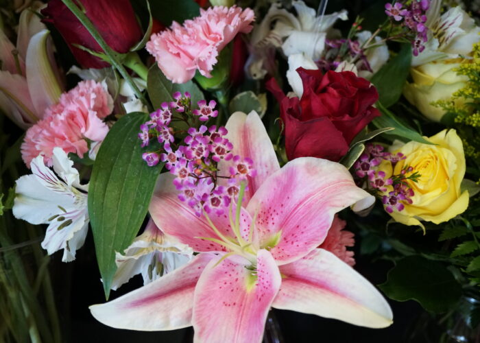 “A vibrant wedding bouquet composed of lilies, roses, mums, alstroemeria, and small waxflowers, arranged in a lush, rounded style with soft greenery.”
