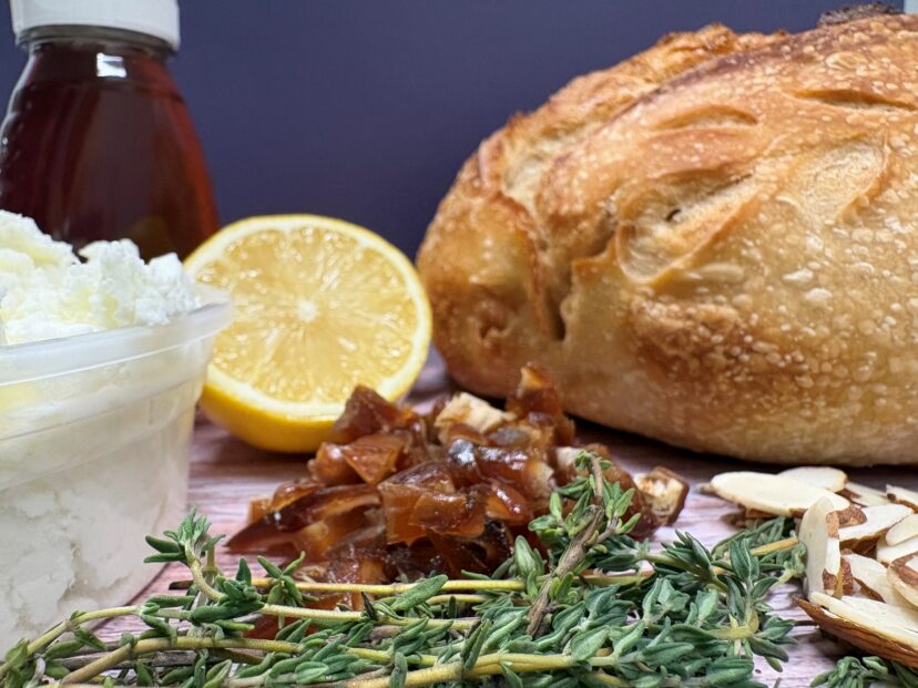 Loaf of bread with feta cheese, lemon, chopped dates, almonds, honey, and fresh thyme on a wooden surface.