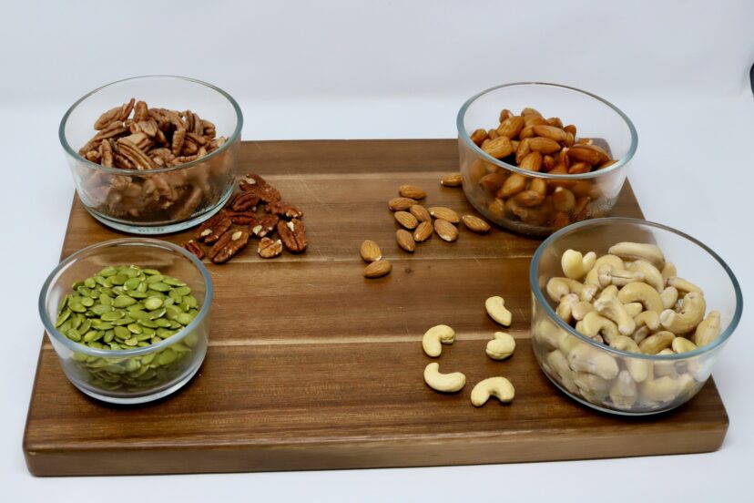 Glass bowls of pecans, almonds, cashews, and pumpkin seeds arranged on a wooden board for making plant-based milk at home.