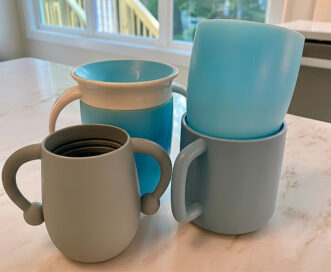 Four light blue and gray toddler cups with handles are arranged on a marble countertop in a bright kitchen.