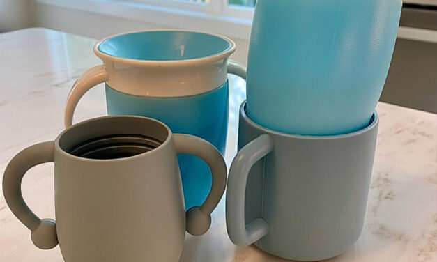 Four light blue and gray toddler cups with handles are arranged on a marble countertop in a bright kitchen.