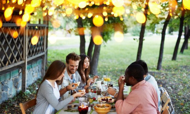Group of people enjoying a picnic outdoors under shade while sharing food at a table.