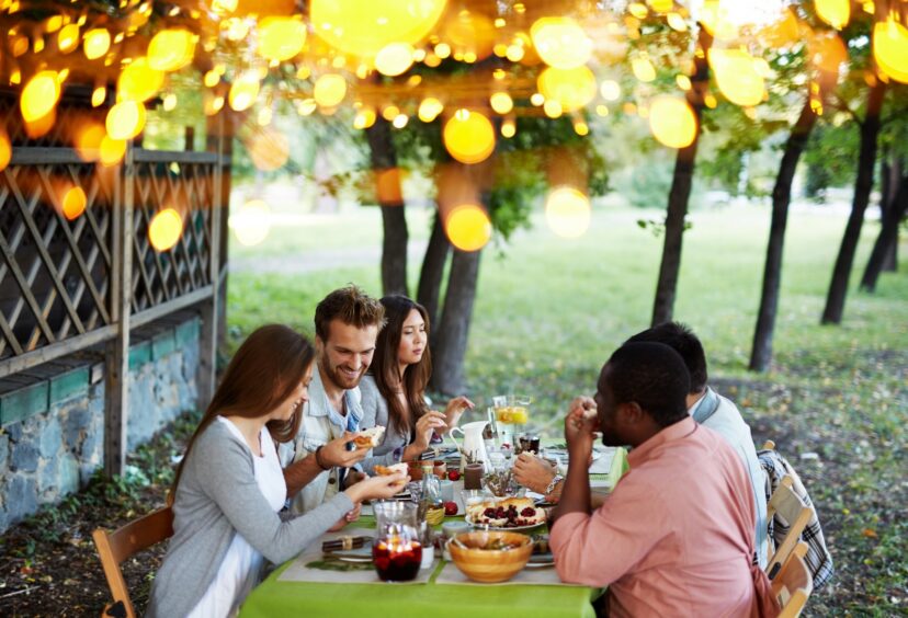 Group of people enjoying a picnic outdoors under shade while sharing food at a table.