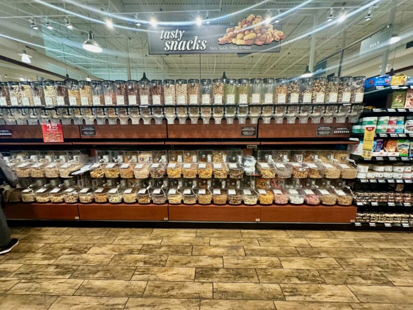 A grocery store bulk food section with dispensers and bins filled with nuts, grains, seeds, spices, dried fruits, and snack mixes.