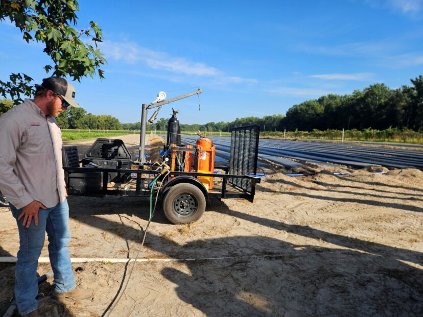A man stands beside a trailer-mounted agricultural fumigation rig parked at the end of plastic-covered strawberry beds.
