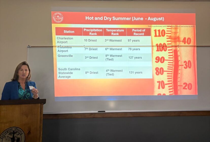 A presenter stands beside a projected slide about hot and dry summer conditions, with a large thermometer graphic.