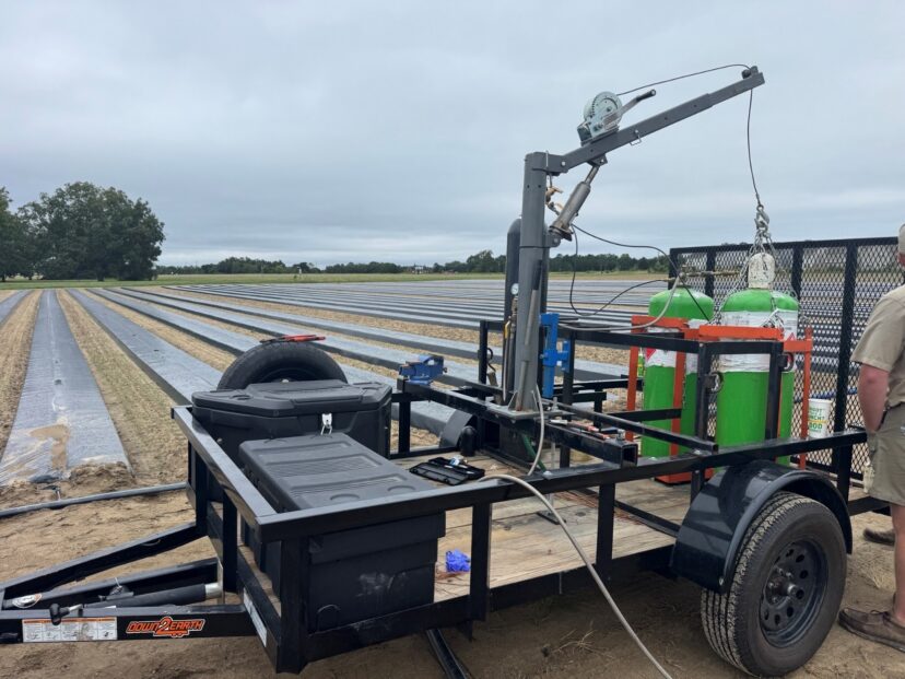 A trailer-mounted fumigation rig with tanks and hoses positioned beside plastic-covered field beds.